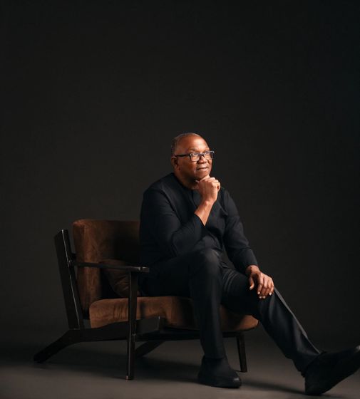 Peter Obi seated in a low leather armchair against a dark ground, in a black tunic, chin resting on one hand, one leg crossed over the other