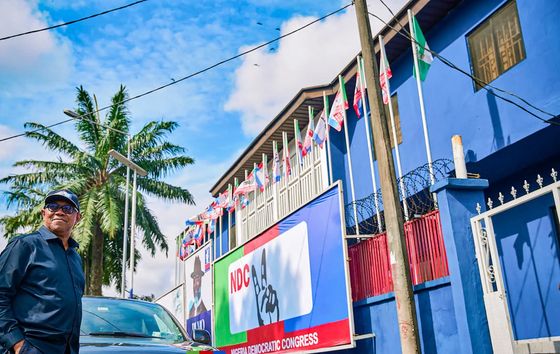 Peter Obi standing beside a vehicle on a street of blue buildings lined with flags and palms