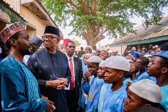 Peter Obi standing among schoolboys in blue uniforms and white caps under a tree in a northern town