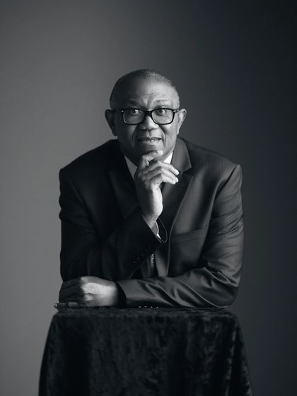 Black and white studio portrait of Peter Obi in a dark suit, leaning on a draped table with his chin resting on one hand