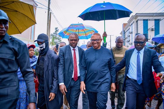 Peter Obi walking down a wet street with officials under blue and yellow umbrellas