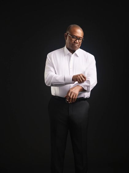Studio portrait of Peter Obi against a black ground in a white shirt and dark trousers, head down, turning back a cuff