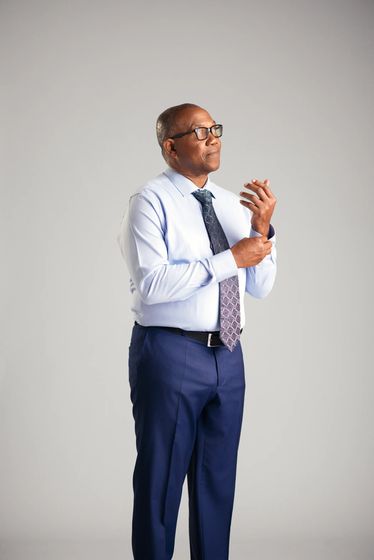 Studio portrait of Peter Obi standing against a pale grey ground in a light blue shirt, patterned tie and navy trousers, fastening a cuff and looking past the camera